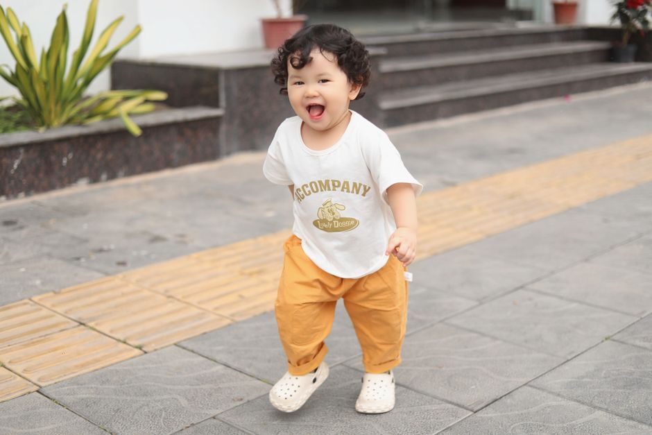 Charming toddler in yellow pants smiles while walking on the pavement in Bắc Giang, Vietnam.